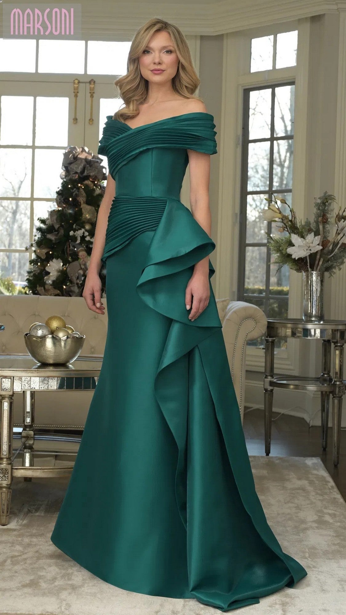 A woman stands indoors in the emerald Long Formal Dress: Marsoni MV1354, an off-shoulder mermaid gown with ruffled details, posed before a decorated Christmas tree and large windows.